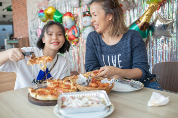 mother and daughter cutting slice of pizza and eating italian pizza together while having enjoying with party to celebrate for weekend in kitchen at home. single mother eats happily with her baby.