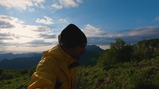 Tourist in yellow windbreaker and buff walking in the mountains with sunset in the background. A man looks at the camera and talks about the hike. Beautiful sunset sky