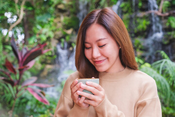 Portrait image of a young woman holding and drinking coffee while sitting in the garden with waterfall