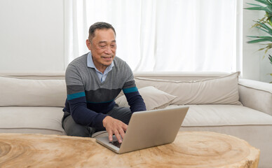 Home office scenario capturing senior man comfortably sitting on a couch, busy working or surfing the internet on his laptop.