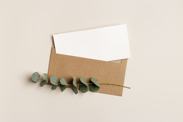 Blank paper card mock up and craft envelope with branches eucalyptus leaves on beige background. White empty invitation on table. Minimal lifestyle photo. Flat lay, top view, copy space.