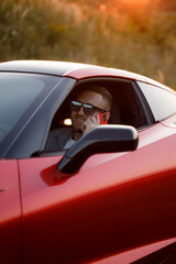 A young handsome man of Caucasian appearance is sitting in a red car outdoors, talking on the phone, against the background of the sunset.
