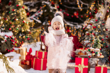 Fototapeta na wymiar A little girl in winter clothes joyfully throws snow into the sky against the backdrop of a Christmas tree and a pile of boxes with gifts in beautiful red packaging. Christmas background.