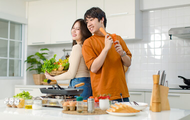Asian young lover couple husband and wife in casual outfit standing smiling posing holding carrots and mixed vegetable basket helping cooking in full decoration modern kitchen with equipment at home