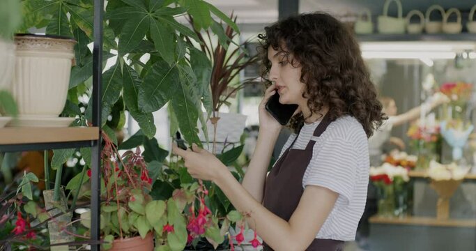 Flower shop employee talking on mobile phone and checking green plants working in store. Modern technology and business communication concept.