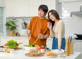 Asian young lovely lover couple husband and wife in casual outfit standing smiling holding red tomato while mixing vegetable salad with wooden fork and spoon in bowl in full decorated modern kitchen