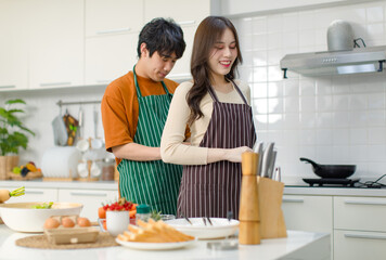 Asian young lovely lover couple husband and wife in casual outfit standing smiling male helping wearing clean striped apron to female in full decorated modern kitchen with cooking equipment at home