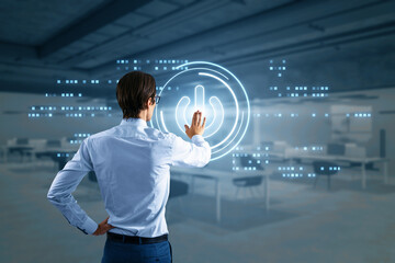 Back view of businessman hand using glowing digital power button on blurry office interior background. Computer and technology concept.