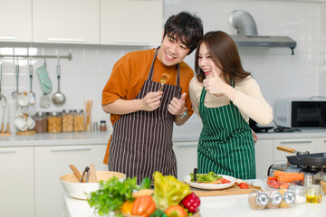 Asian young lover couple husband and wife in casual outfit with apron standing smiling woman using fork feeding delicious steak to man in full decorated modern kitchen with ingredients and equipment
