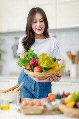 Asian young beautiful housewife in casual outfit standing smiling showing red sweet pepper and mixed fresh organic vegetables basket in full decorated modern kitchen with ingredients and equipment