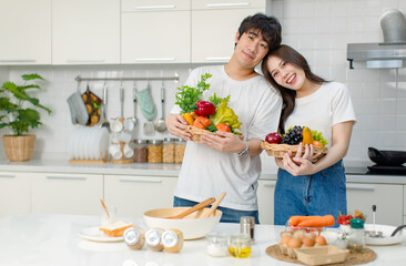 Obraz na płótnie Canvas Asian young lovely lover couple husband and housewife in casual outfit standing smiling posing holding mixed fruits and vegetables basket in full decorated modern kitchen with ingredients equipment