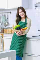 Asian young beautiful housewife in casual outfit with apron standing smiling holding mixed fresh organic fruits basket and vegetables basket in full decorated modern kitchen with ingredients at home