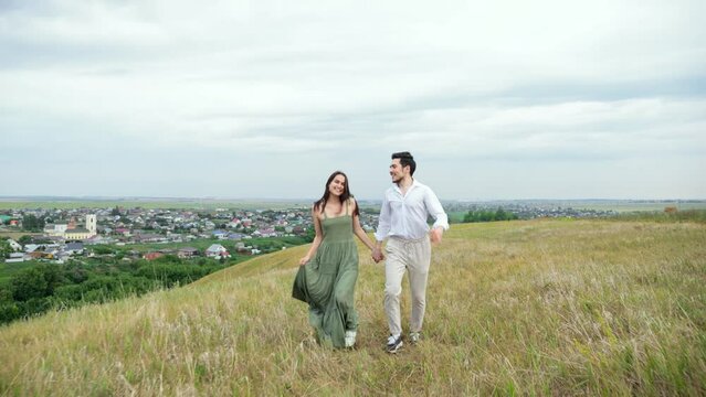 Positive couple of man and woman run across hilly field against huge town. Concept of view of territory with buildings and love slow motion