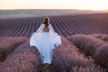 Happy woman in a white dress and straw hat strolling through a lavender field at sunrise, taking in...