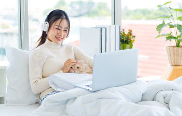 Asian pretty female teenager girl in turtleneck sweater wearing headphones listen to music from laptop on white clean sheet bed under blanket drinking coffee with cute domestic tabby short hair cat