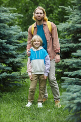 Fototapeta premium Vertical full length portrait of modern father and son looking at camera together standing in forest
