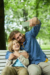 Portrait of father and son sitting on bench in park together and taking selfie photo