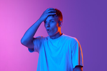 Young man holding hand on head and standing with shocked face against gradient pink purple background in neon light. Concept of human emotions, youth, lifestyle, fashion, facial expressions, ad