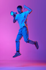Emotional young man shouting in megaphone against gradient pink purple background in neon light. News. Concept of human emotions, youth, lifestyle, fashion, business, mass media, ad