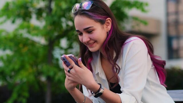 Beautiful young girl receiving good news on her mobile phone while sitting on the bench outdoor. Surprised female student smiling happily looking at the smartphone. Woman in love.