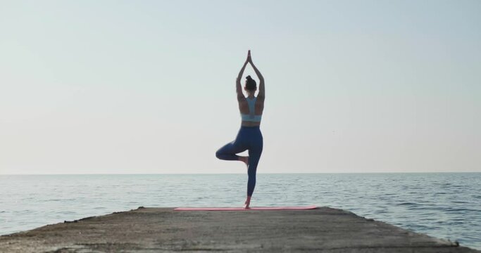 Sporty young woman practicing yoga on pier in morning, back view