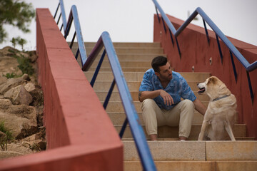 Young Hispanic man, sitting on stairs next to his dog while talking with him in a loving and tender attitude. Concept, dogs, pets, animals, friends.