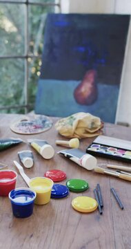 Vertical video of close up of tubes and tubs of paint and brushes on table in studio, slow motion