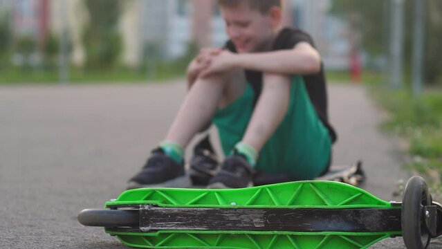 a boy of 8-9 years old in defocus sitting on the pavement after falling from a scooter stroking his sore knee. accident, fall from a bicycle, scooter. medical insurance for children