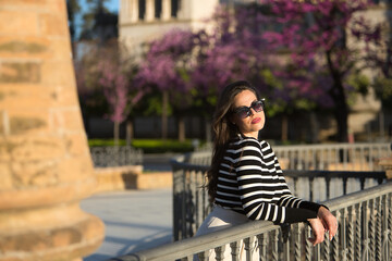 Obraz premium Young and beautiful woman, Hispanic, brown hair, with striped sweater, linen pants and sunglasses, leaning on a railing, looking at the camera relaxed and calm. Concept relax, peace, tranquility.