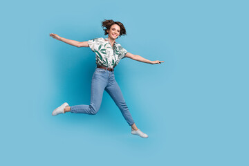 Fototapeta na wymiar Full length photo of sweet carefree lady wear print shirt jumping high flying arms sides empty space isolated blue color background