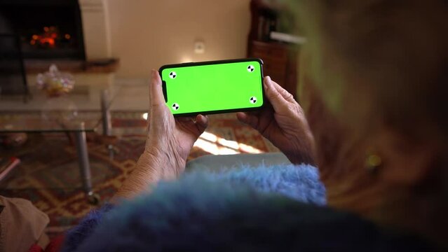Old woman at home using a mobile phone with green screen. Mockup device to add elements. Wrinkled hands of grandmother holds a smartphone. Concept of technology
