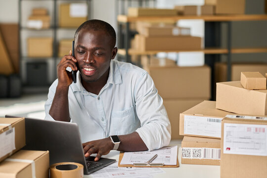 Young African American man in white shirt sitting by workplace in warehouse or marketplace and talking on mobile phone to client