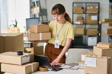 Young woman in casualwear holding stack of packed boxes and using laptop while checking personal...