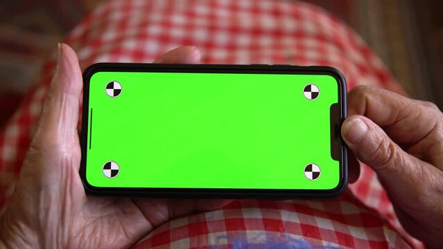 Old woman at home using a mobile phone with green screen. Mockup device to add elements. Wrinkled hands of grandmother holds a smartphone. Concept of technology