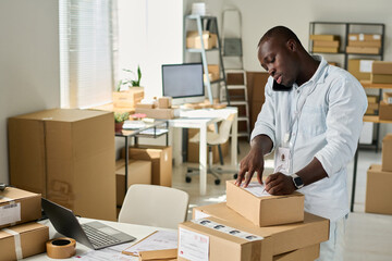 African American manager of marketplace filling in personal data of customer on top of packed box while talking to client on smartphone