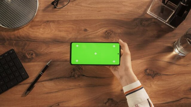 Anonymous Person Using a Smartphone with Mock Up Green Screen Chromakey Display with Placeholders. Person Holding Cellphone Vertically, Watching Videos on a Wooden Table Background. Top Down View