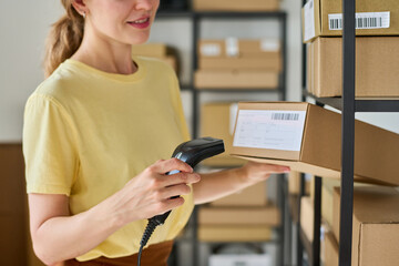 Cropped shot of young female worker of warehouse scanning qr codes on boxes while standing in front of shelves with packed online orders