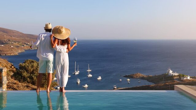 A hugging, loving couple on vacation time enjoys the summer sunset over the Aegean Sea by the swimming pool with a drink