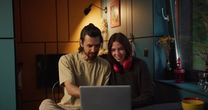 A brunette guy and a brunette girl in red headphones sit and watch a video on a laptop in the evening with a yellow lamp on the background
