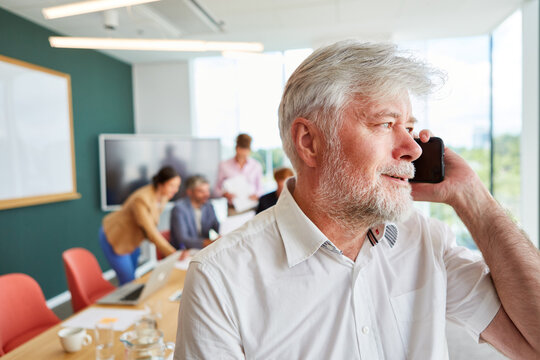 Bearded senior businessman talking on mobile phone while sitting in front of colleagues discussing at office