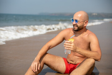 Smiling vacationist in sunglasses relaxing on sandy beach with drink decorated with pineapple in hand, looking at sea.