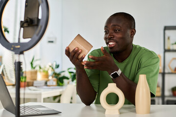 Happy young craftsman or designer showing online audience handmade vases while sitting in front of...