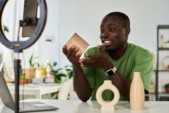 Happy young craftsman or designer showing online audience handmade vases while sitting in front of smartphone screen during livestream