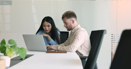 Asian and Caucasian colleagues using laptop sit at desk in office, preparing presentation, discuss corporate software, mentor helps to apprentice with new program. Workflow, teamwork use modern tech