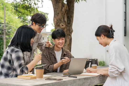 Group of happy young Asian college students sitting on a on the cement table, looking at a laptop screen, discussing and brainstorming on their university project together - Powered by Adobe