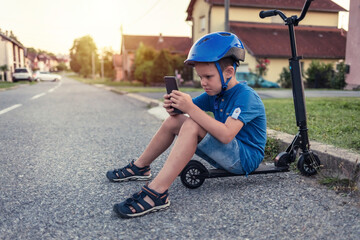 Obraz premium Lonely boy playing games on his smartphone, sitting on his push scooter during the day.