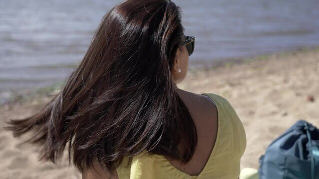 Slow motion video of a girl sitting on the beach. The girl straightens her hair, which develops the sea breeze. Beautiful woman on vacation.