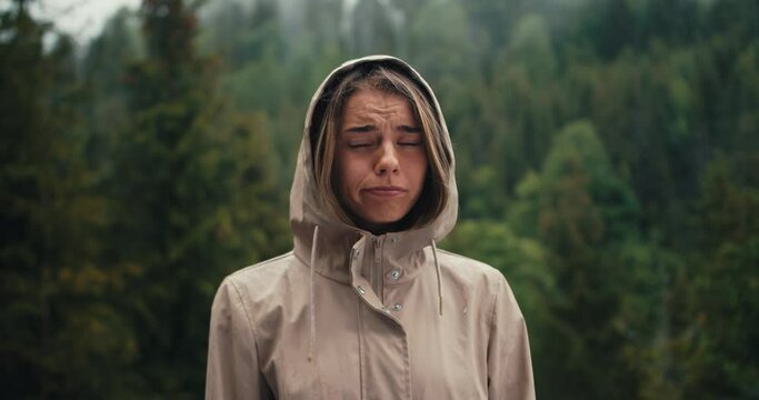 A sad blonde girl stands against the background of forest rain in the mountains, she is sad and looks at the camera