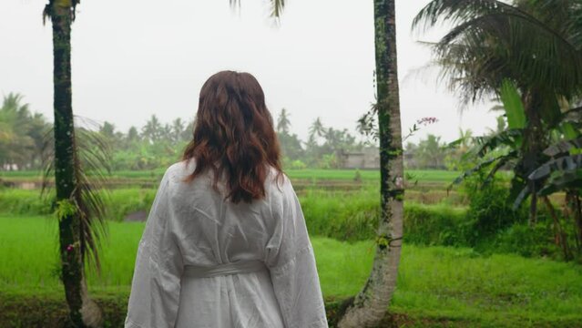 Back view of woman in nightgown standing and looking at green fields with grass, tropical palm trees and sky. Panoramic window of Bali beautiful nature. Concept of holiday and top destination