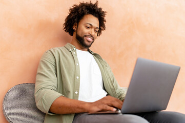 Positive young african-american man sitting in the loft style office and using laptop. Cheerful male employee typing on the keyboard, looks at the screen satisfied